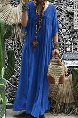A woman wearing a long, flowing royal blue maxi dress with short sleeves and a v-neckline. She is accessorized with a beaded necklace and bracelets and is carrying a woven bag. The background features cacti and decorative wall art.
