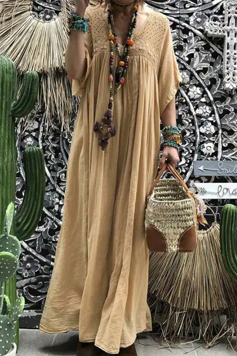 A woman wearing a long, flowing tan maxi dress with short sleeves and a v-neckline. She is accessorized with a beaded necklace and bracelets and is carrying a woven bag. The background features cacti and decorative wall art.