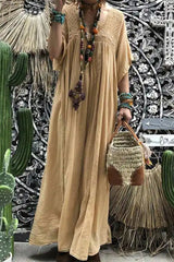 A woman wearing a long, flowing tan maxi dress with short sleeves and a v-neckline. She is accessorized with a beaded necklace and bracelets and is carrying a woven bag. The background features cacti and decorative wall art.