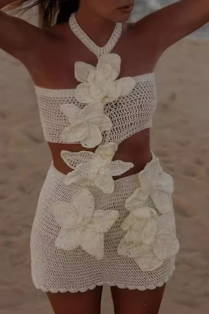 A woman in a white crochet bikini set with large white floral appliqués, standing on a sandy beach.