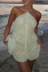A woman poses on a beach at sunset wearing a cream-colored, heavily beaded mini dress with a halter neckline and fringe detailing on the hem and a matching clutch.