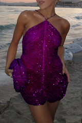 A woman poses on a beach at sunset wearing a vibrant purple, heavily beaded mini dress with a halter neckline and fringe detailing on the hem and a matching clutch.