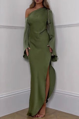 Full-length shot of a woman in an olive green satin gown with an asymmetrical neckline and long, flared sleeves. The dress has a high slit, and she is accessorized with a gold chain bag.