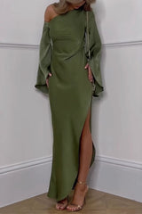 A woman stands in a corner wearing a flowing olive green satin dress with a one-shoulder design and dramatic bell sleeves. The dress features a high slit on the side, revealing her leg, and she is wearing gold strappy heels.