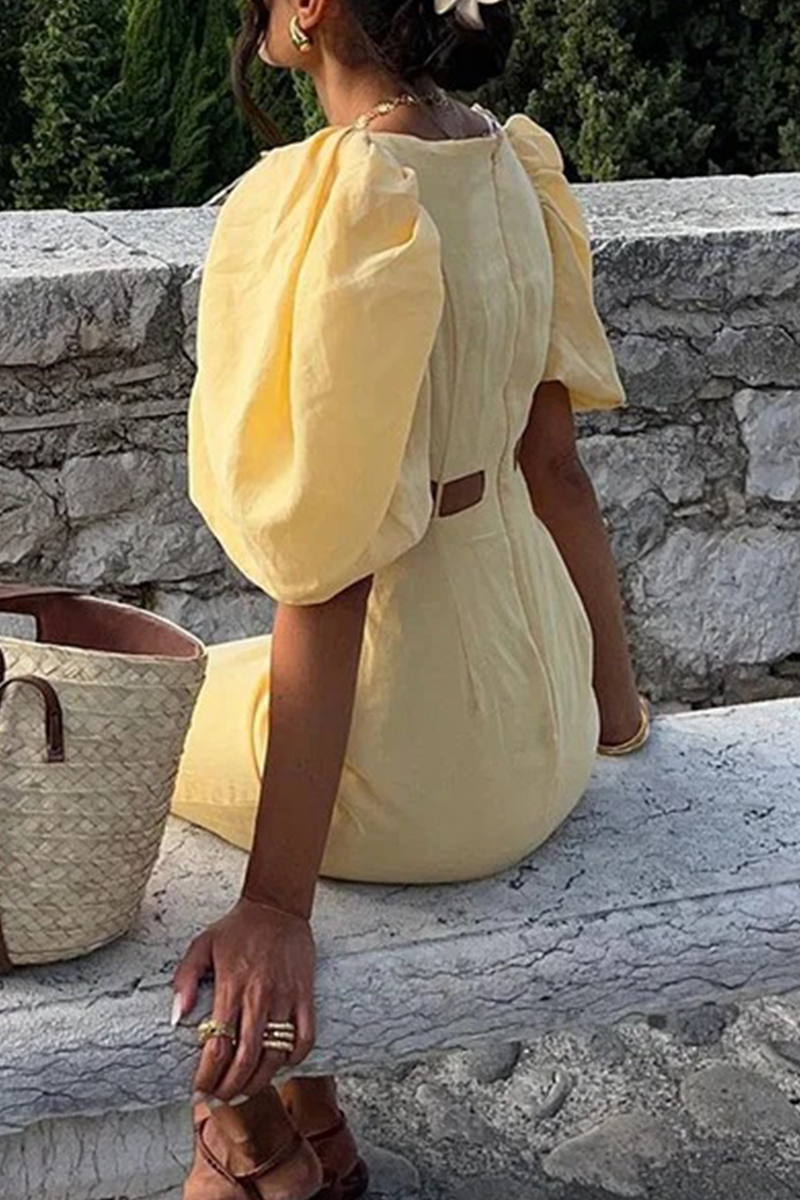 A woman wearing a yellow linen dress with puff sleeves and a cut-out detail, sitting on a chair and reading.