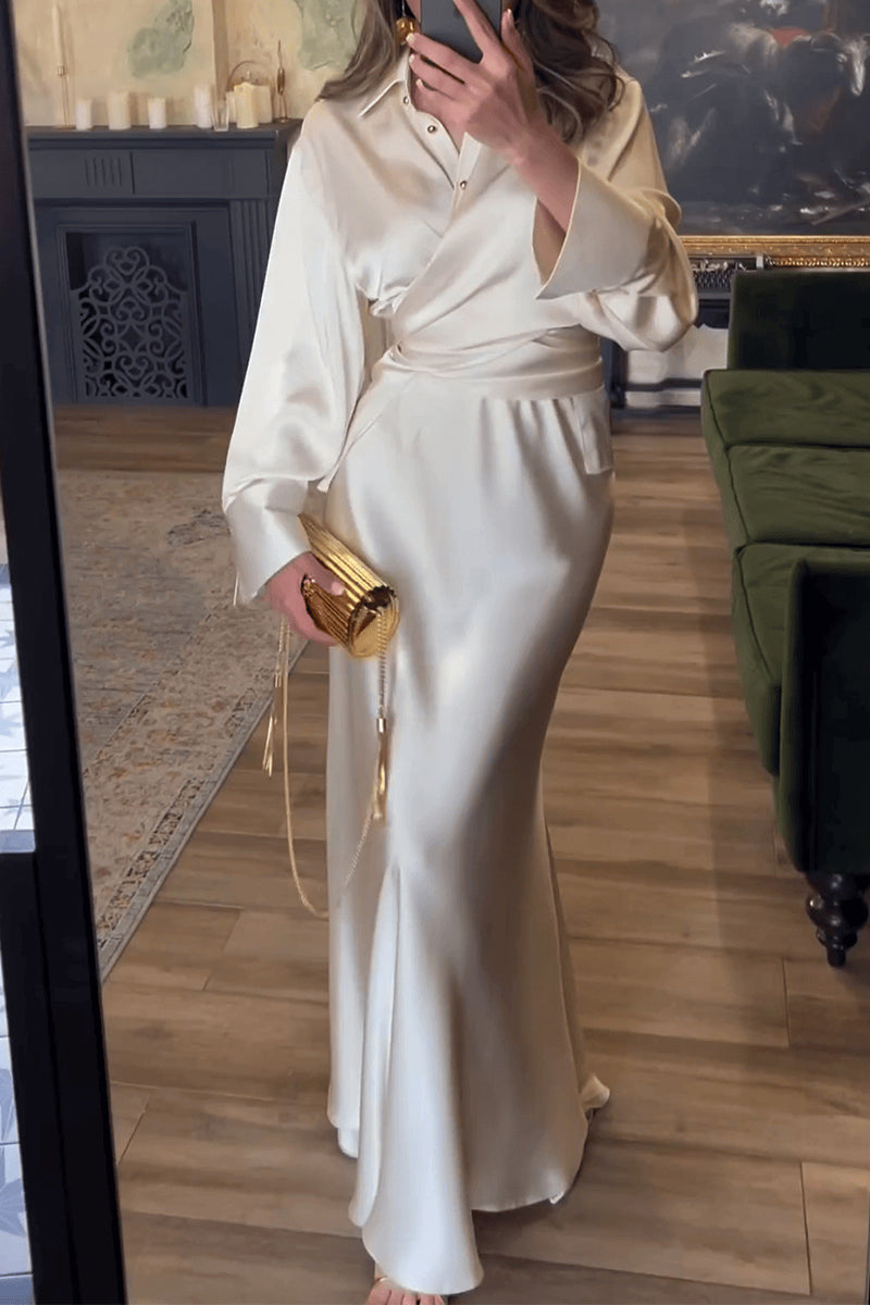 Woman in a cream satin wrap dress with a belted waist and a gold clutch bag, standing in front of a mirror.