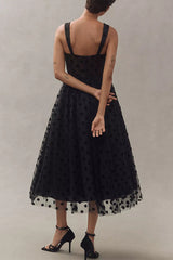 Back view of a woman wearing a black polka dot tulle midi dress with a sweetheart neckline and spaghetti straps, standing with her arms crossed.