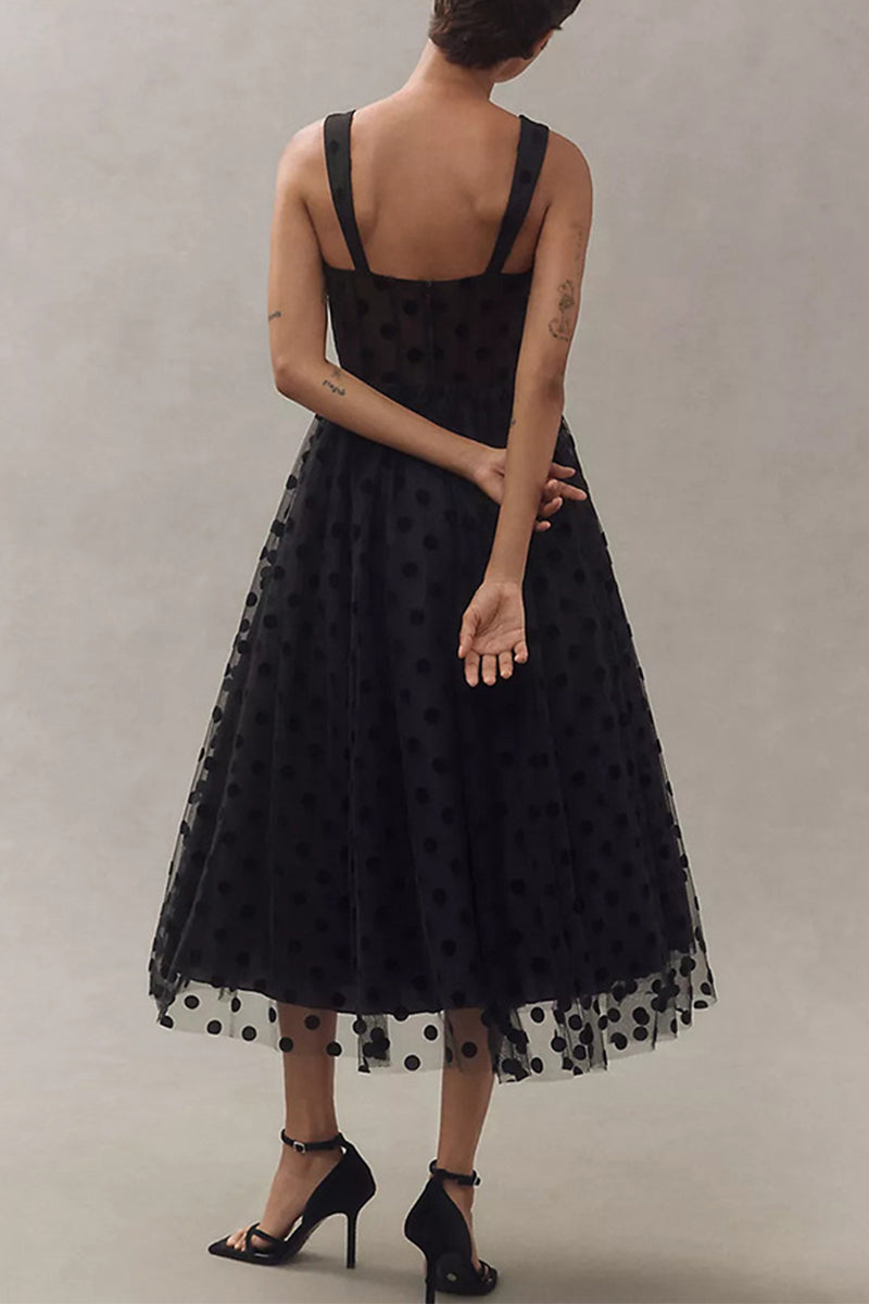 Back view of a woman wearing a black polka dot tulle midi dress with a sweetheart neckline and spaghetti straps, standing with her arms crossed.