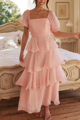 Woman in a pink tiered ruffle dress with puff sleeves smiling and posing by a bed.