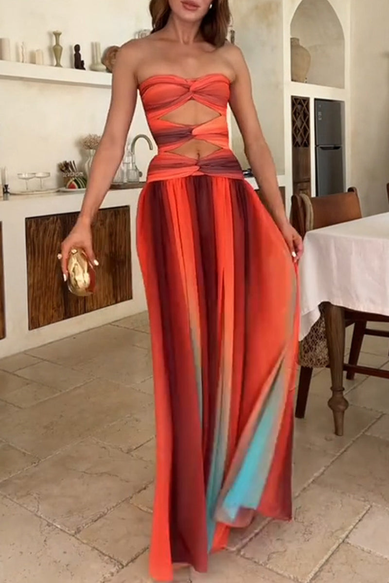 A woman in a strapless orange, red, and blue ombre maxi dress with cut-outs and a twist-front bodice, holding a gold clutch, walking in a modern interior.