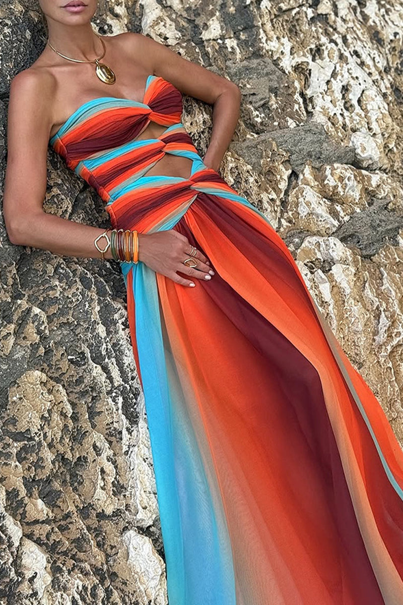A woman in a strapless orange, red, and blue ombre maxi dress with cut-outs and a twist-front bodice, leaning against a rock formation, accessorized with bracelets and rings.