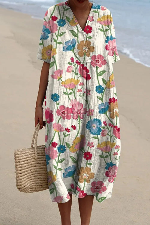 A woman wearing a white midi dress with a bold floral print in pink, blue, and yellow, v-neck, and short sleeves, holding a straw tote bag on a beach.
