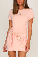 A woman wearing a pink short-sleeve crew neck dress with a tie waist and ruched detailing.
