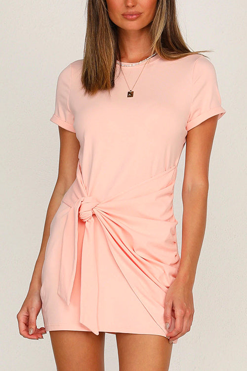 A woman wearing a pink short-sleeve crew neck dress with a tie waist and ruched detailing.