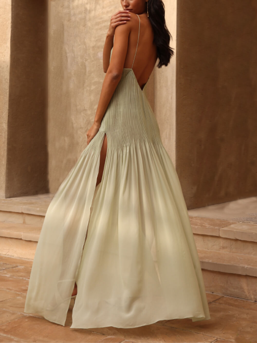 back view of a woman in a pale green pleated maxi dress with thin straps and a high slit, showing her back