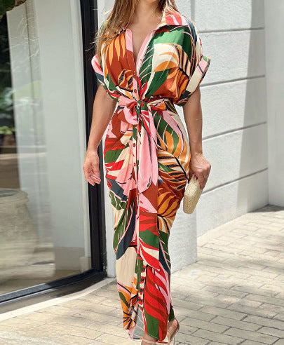 Woman wearing a long, flowing dress with a vibrant tropical leaf print in shades of green, orange, red, and pink.
