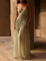 woman in pale green pleated maxi dress with thin straps and a deep V-neckline, standing in an outdoor setting with beige walls