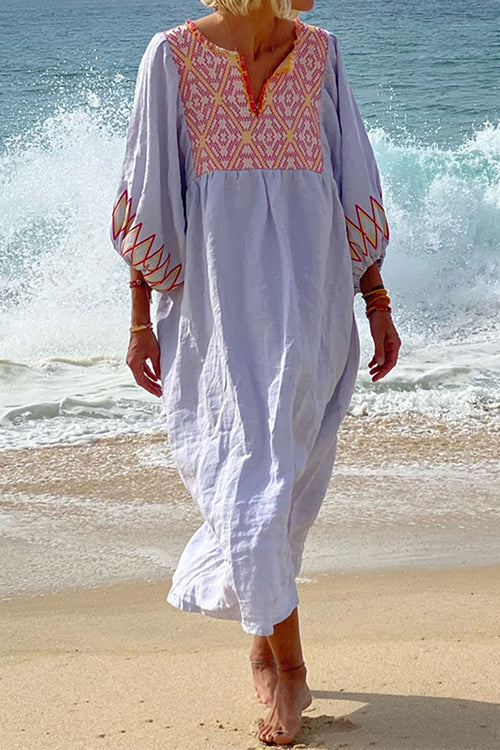Woman in a light blue maxi dress with pink embroidery on the neckline and sleeves, walking on a beach with a wave crashing behind her.