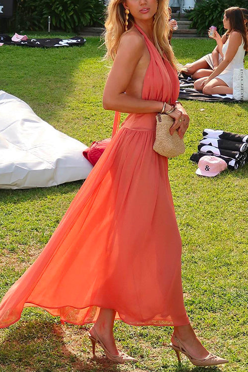Woman wearing a coral halter neck maxi dress with a flowy skirt and a small straw bag, posing outdoors on a grassy lawn.