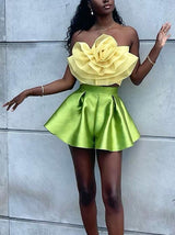 A full-body shot of a woman in a yellow floral strapless top and green pleated shorts, standing against a wall.