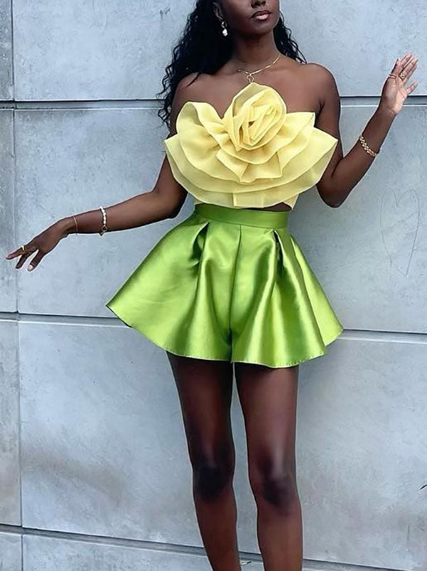 A full-body shot of a woman in a yellow floral strapless top and green pleated shorts, standing against a wall.