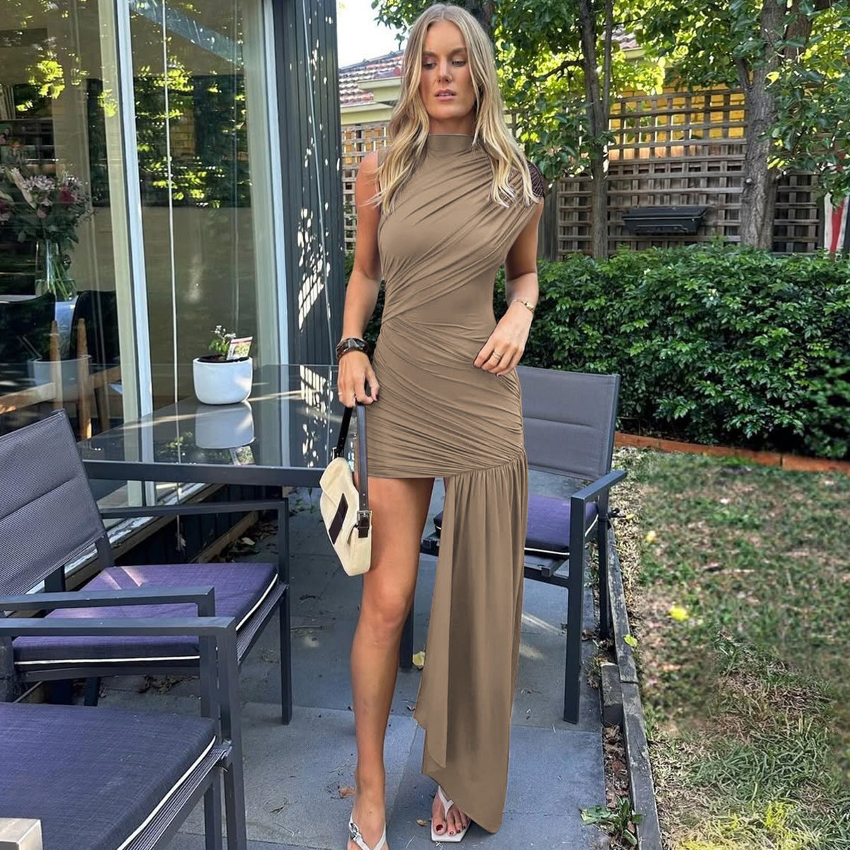 A woman in a taupe ruched dress with an asymmetrical hemline stands outdoors.