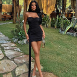 A woman wearing a black strapless ruched mini dress with a gold handbag and silver heels, posing in a garden.
