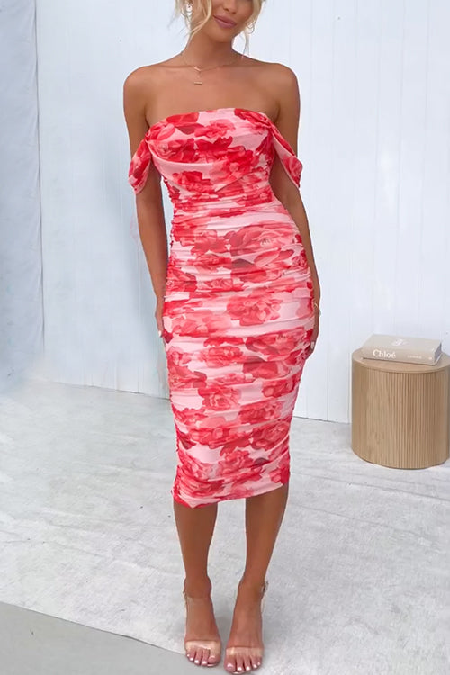 A woman stands in a bright room wearing a red floral print, off-the-shoulder, ruched midi dress and pink heels.