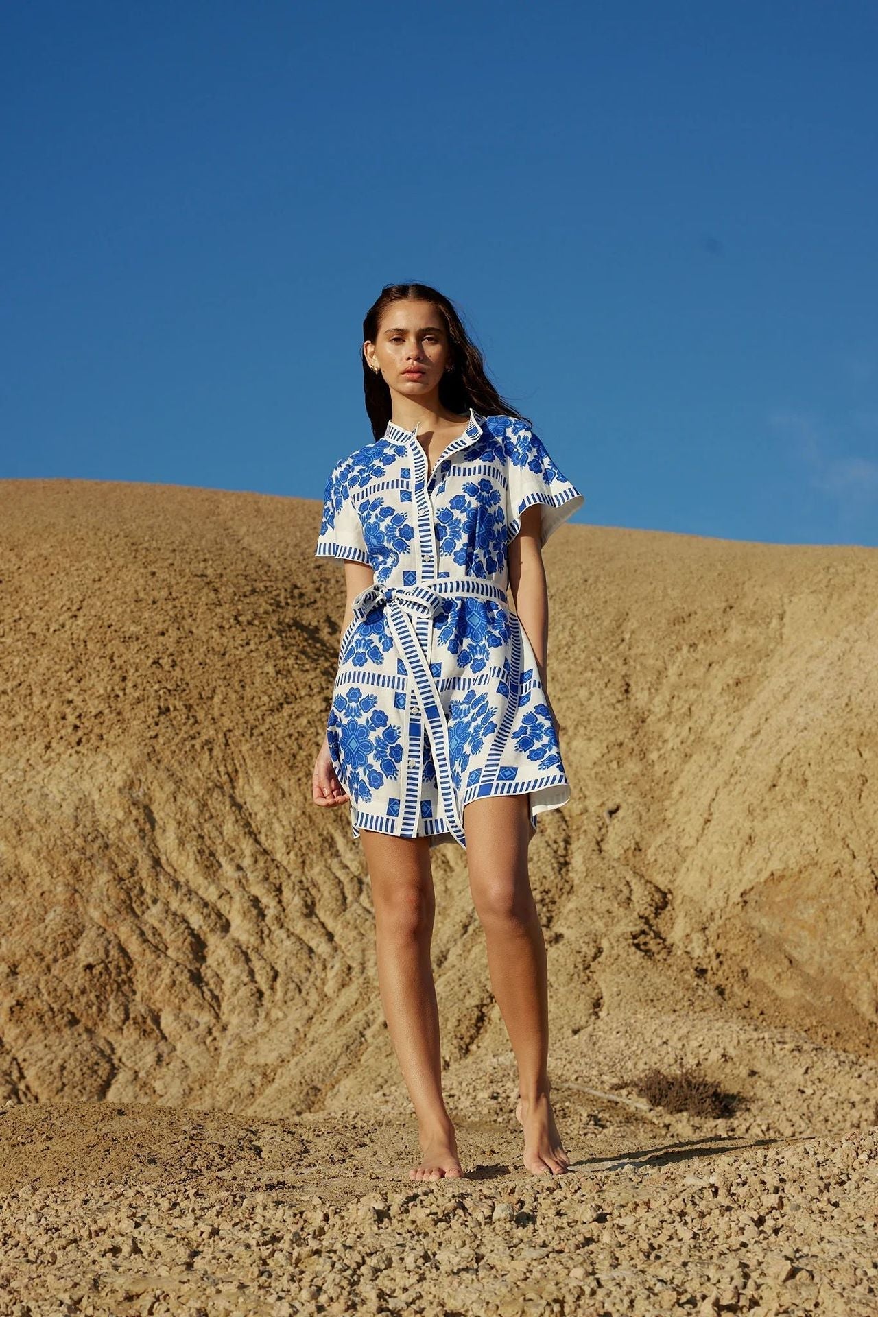 A woman stands in a desert landscape wearing a white and blue floral print shirt dress.