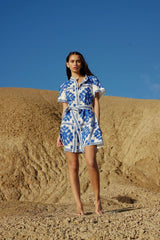 A woman stands in a desert landscape wearing a white and blue floral print shirt dress.