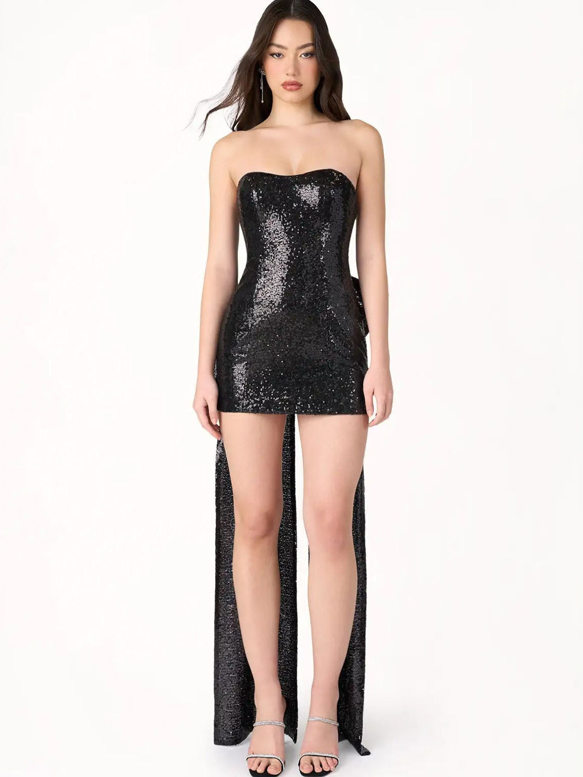 Woman wearing a black sequin strapless mini dress with a long train and a large bow on the back, standing against a white background.