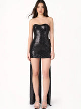 Woman wearing a black sequin strapless mini dress with a long train and a large bow on the back, standing against a white background.