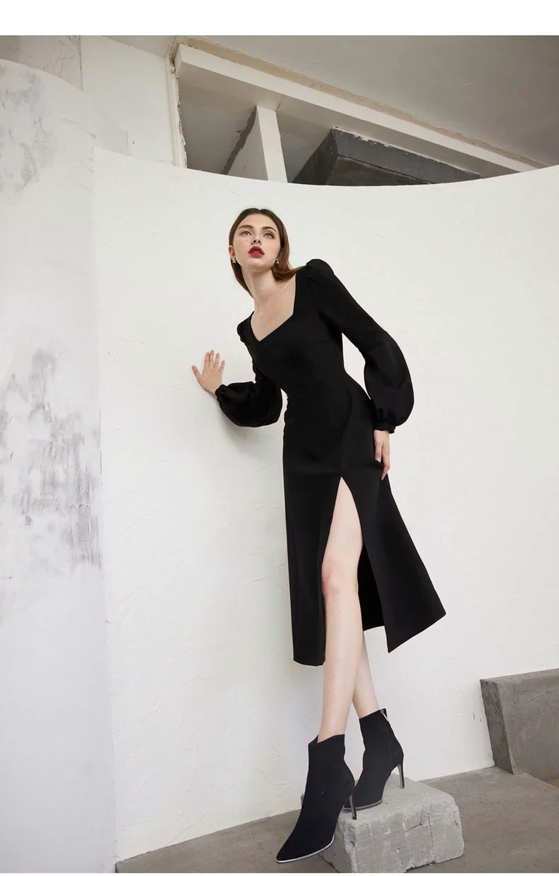 A woman leaning against a white wall in a black midi dress with a square neckline and puff sleeves, with a thigh-high slit.