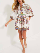 A woman in a white dress with a vibrant paisley print, with her hand on her hip and looking to the side.