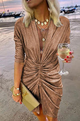 A woman in a shimmering gold ruched dress with a plunging V-neck and three-quarter sleeves, holding a clutch and a glass of white wine on a beach.