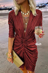 A woman in a shimmering red ruched dress with a plunging V-neck and three-quarter sleeves, holding a clutch and a glass of white wine on a beach.