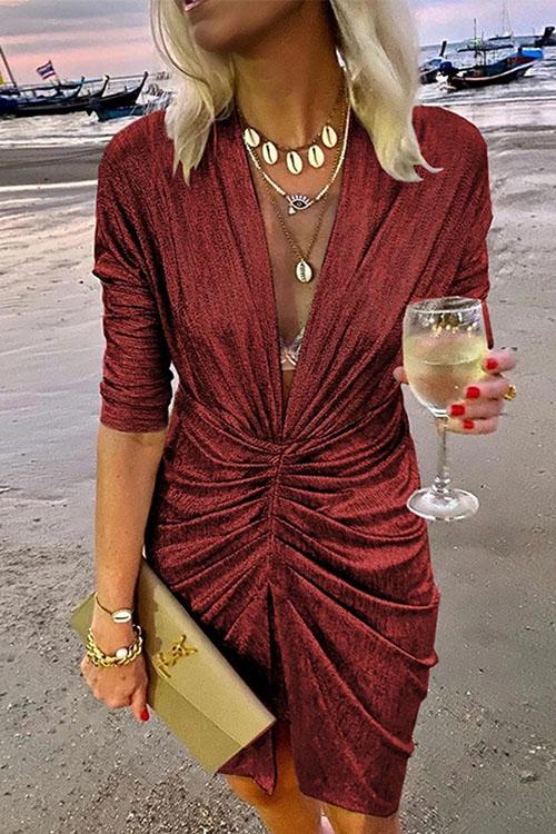 A woman in a shimmering red ruched dress with a plunging V-neck and three-quarter sleeves, holding a clutch and a glass of white wine on a beach.