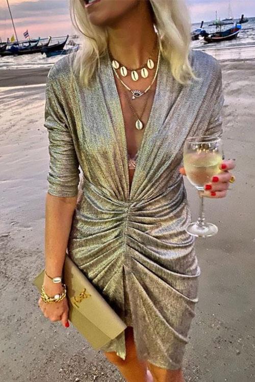A woman in a shimmering silver ruched dress with a plunging V-neck and three-quarter sleeves, holding a clutch and a glass of white wine on a beach.