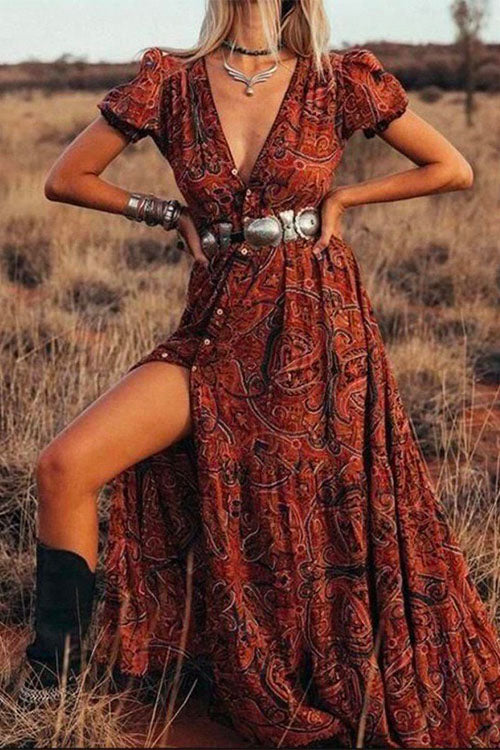 Woman posing in a red paisley maxi dress with a high slit, silver belt, and cowboy boots in a dry field.