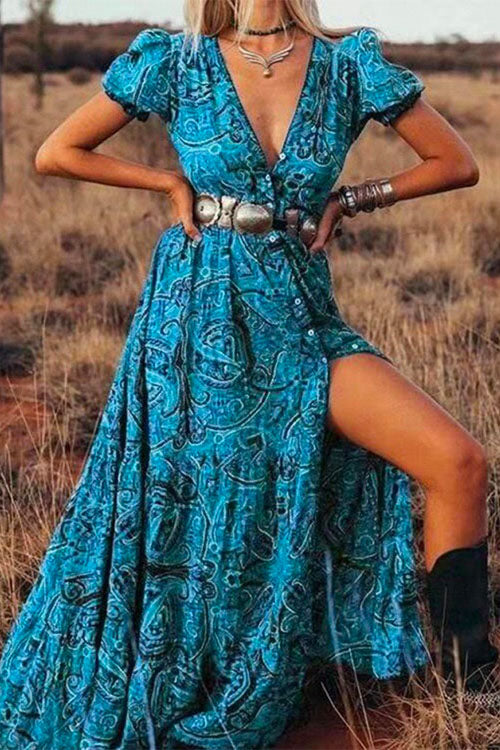 Woman posing in a blue paisley maxi dress with a high slit, silver belt, and cowboy boots in a dry field.