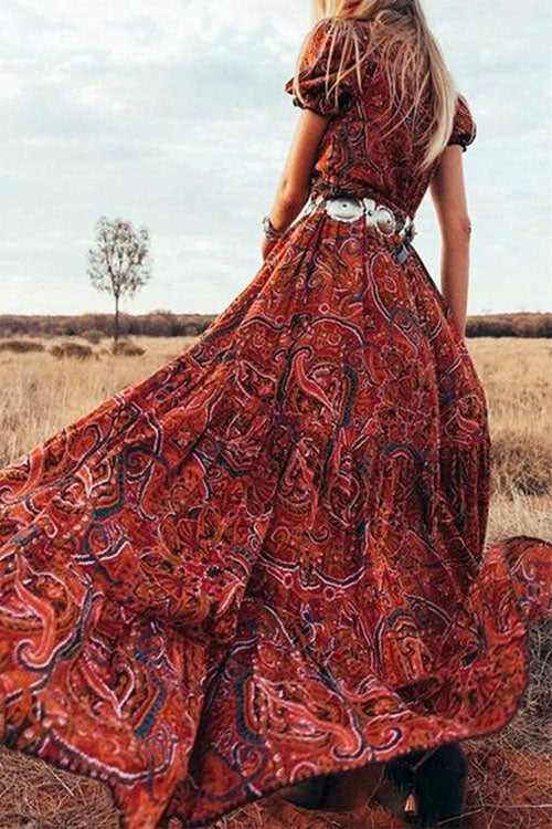 Woman in a flowing red paisley maxi dress with a wide silver belt, standing in a dry, grassy field.