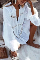Woman wearing a white sheer dress with black embroidered details and ruffled sleeves, sitting on a beach.