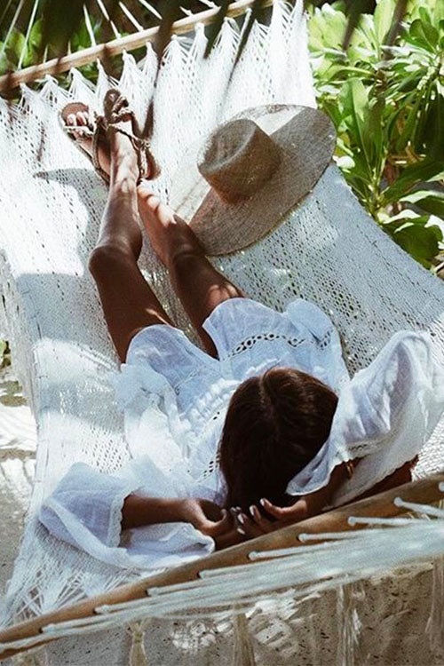 Woman lying in a hammock wearing a white sheer dress with black embroidered details and ruffled sleeves, with a straw hat nearby.