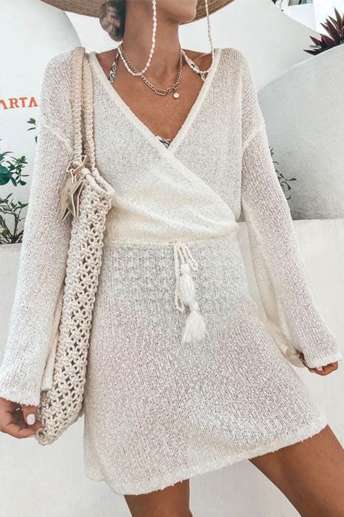 A woman wearing a white knit mini dress with a wrap front and bell sleeves, holding a straw bag.