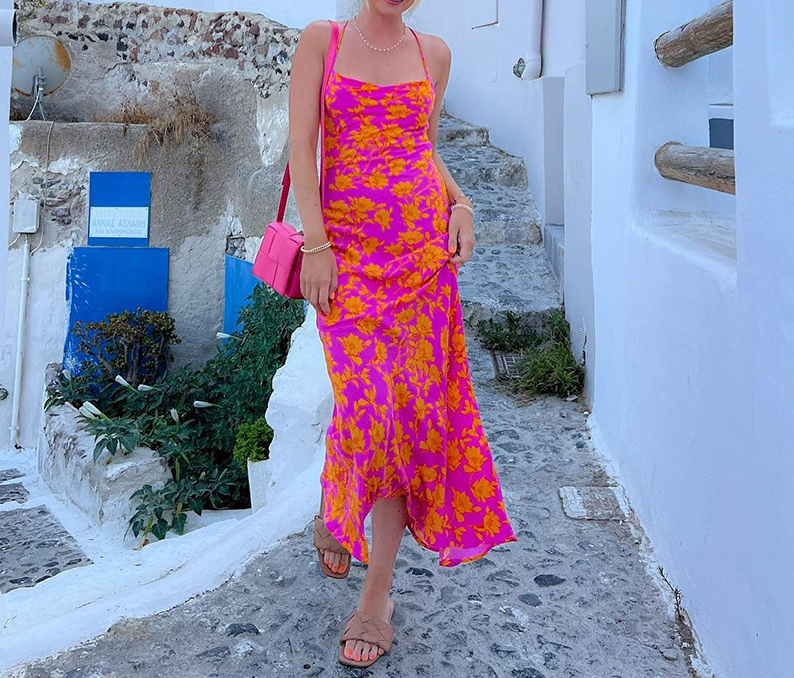 A woman in a pink and orange floral print slip dress stands on a balcony overlooking a white village with a blue sea in the distance.