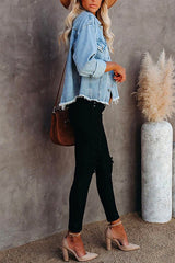 Side view of a woman in a light blue distressed denim jacket, white tank top, black ripped jeans, brown belt, and nude heels, with a tan hat and brown crossbody bag.