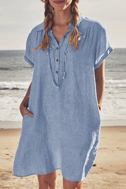 Woman wearing a light blue linen-like short-sleeved collared shirt dress with pockets and a necklace, standing on a beach.