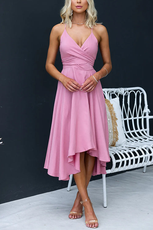 Woman wearing a pink high-low midi dress with a cross-over neckline and tie waist, standing next to a white chair.