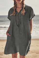 Woman wearing a dark gray linen-like short-sleeved collared shirt dress with pockets and a necklace, standing on a beach.