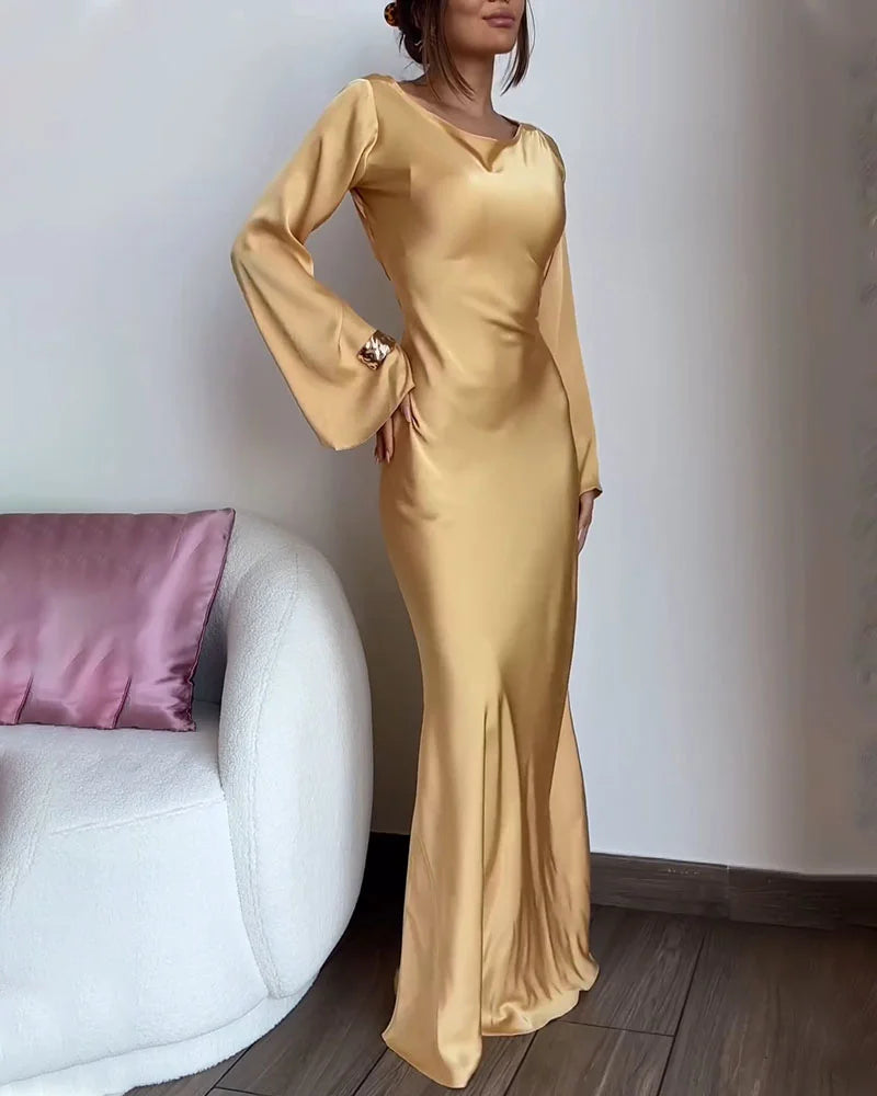 Woman in a gold satin dress with long bell sleeves, standing next to a white sofa.